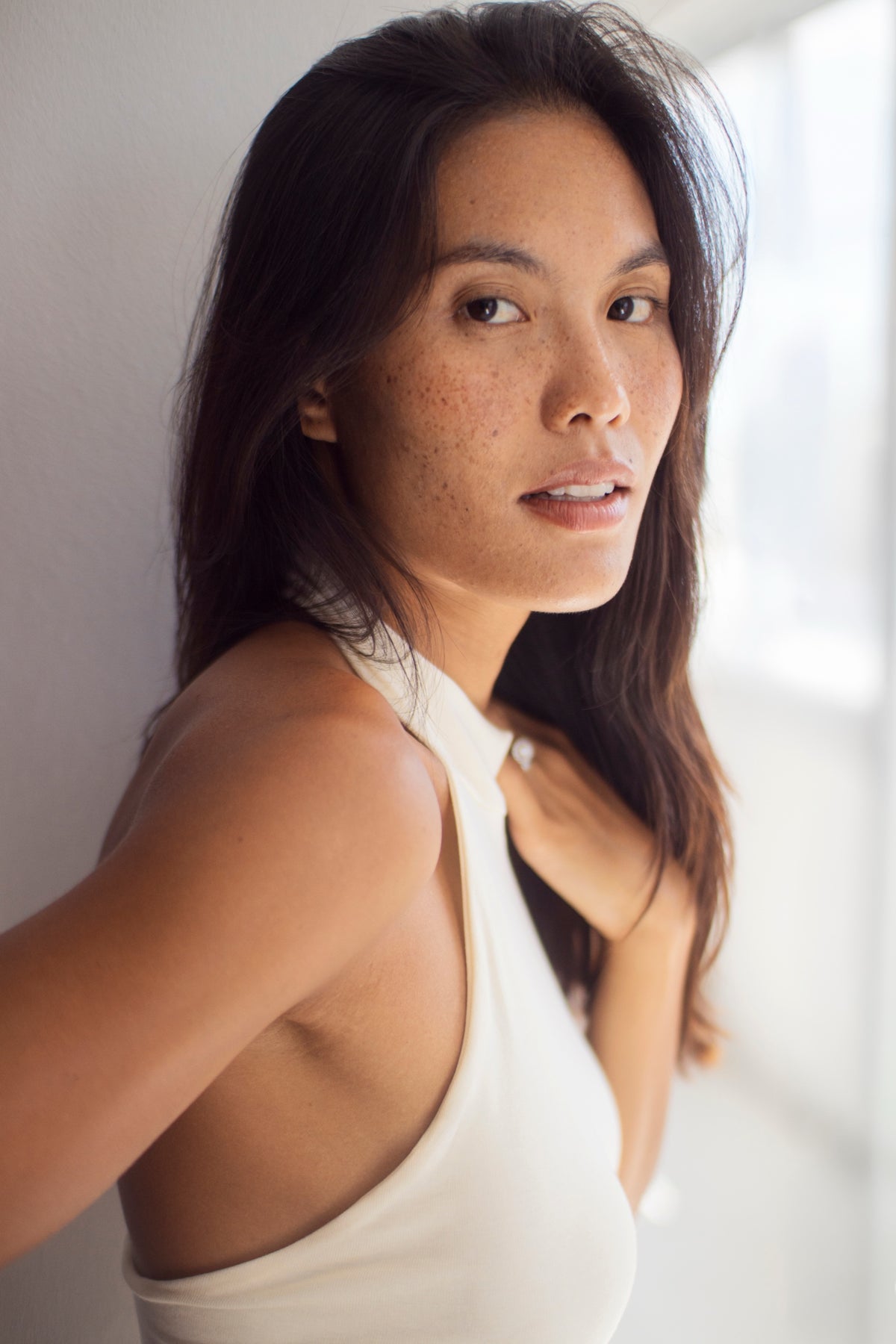 A woman looking over her shoulder beside a light wall.